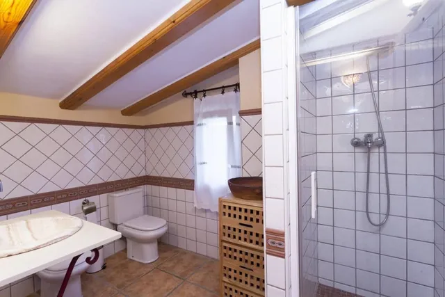 Aseo con plato de ducha y mampara de cristal, inodoro y lavabo, bajo techo abuhardillado con vigas de madera