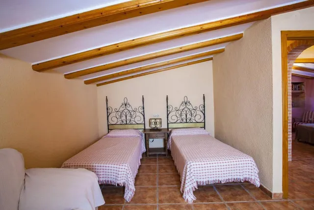 Dormitorio con dos camas individuales, cabeceros de forja calada y colchas de cuadros rojos bajo el techo de vigas de madera