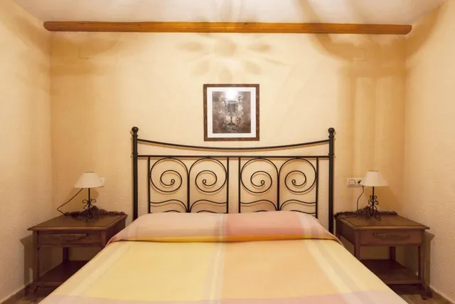 Dormitorio de paredes color crema con cama de matrimonio, cabecero de forja en espiral y dos mesillas de madera con lámparas a juego