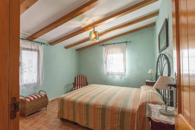 Dormitorio de paredes verde agua con cama de matrimonio, colcha de rayas, butaca y baúl de mimbre bajo el techo abuhardillado