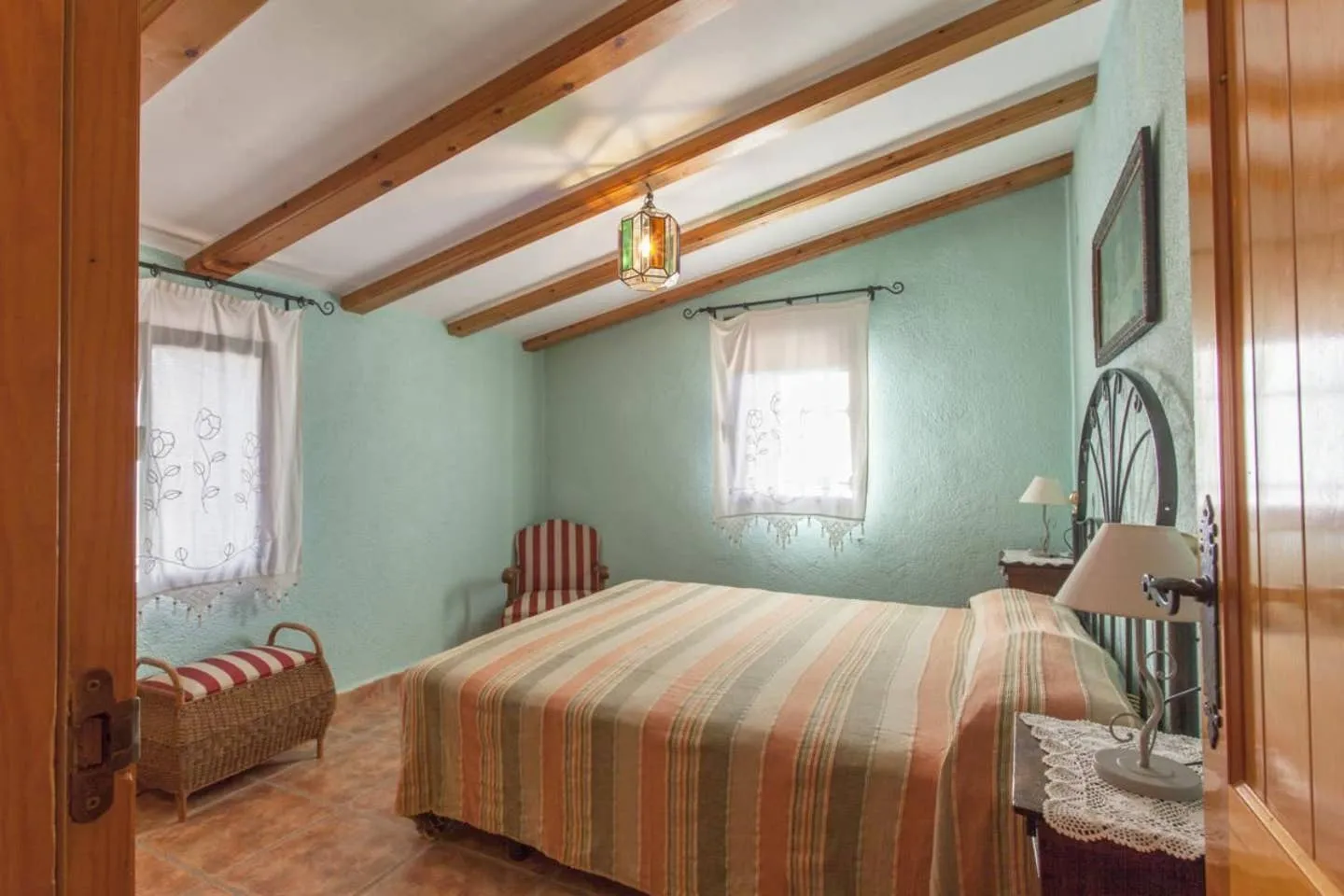 Dormitorio de paredes verde agua con cama de matrimonio, colcha de rayas, butaca y baúl de mimbre bajo el techo abuhardillado