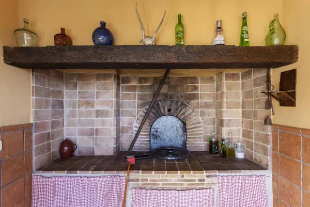 Paellero de obra con hogar de leña, quemador para paella y repisa de madera antigua con botellas de vidrio