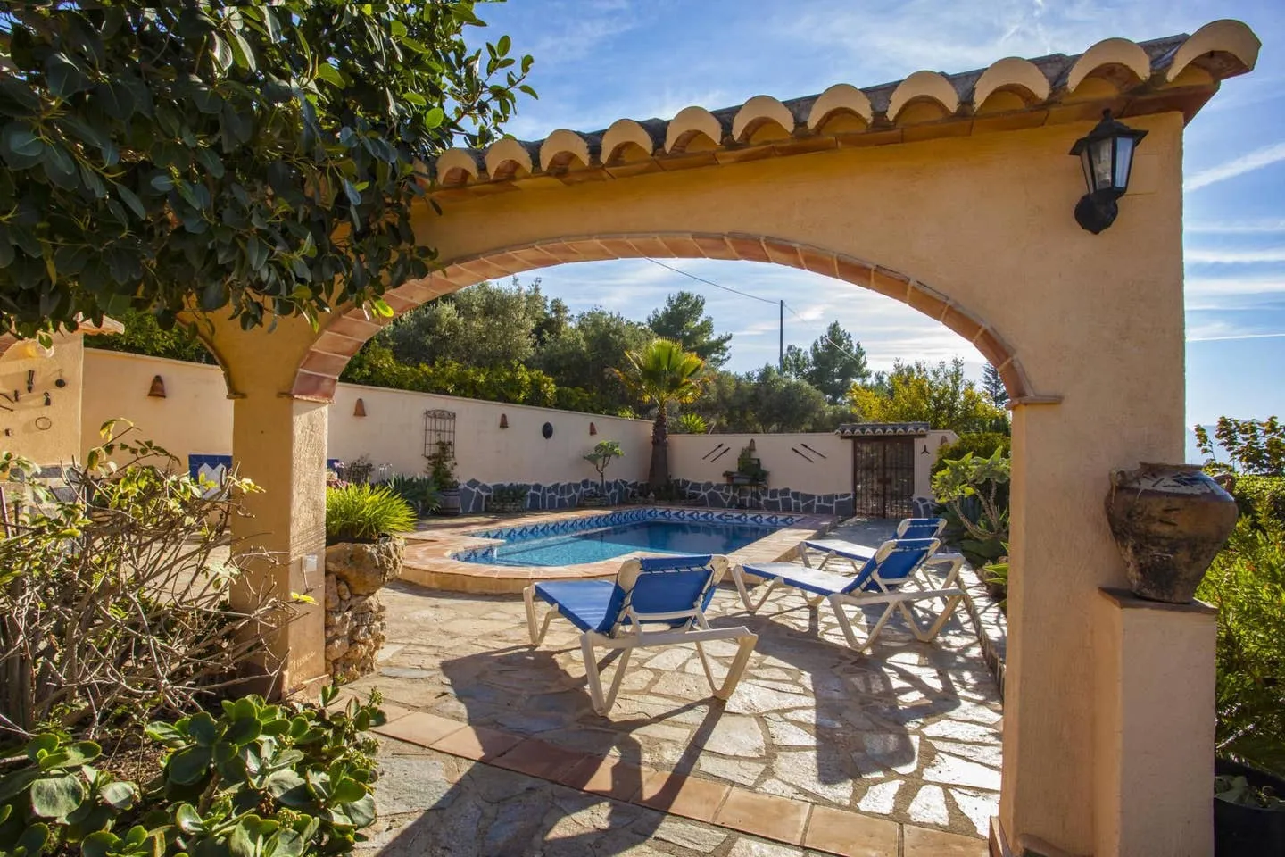 La piscina vista desde el porche, a través de un arco de obra rematado con teja árabe, con dos tumbonas azules al borde del agua