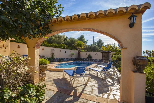 La piscina vista desde el porche, a través de un arco de obra rematado con teja árabe, con dos tumbonas azules al borde del agua