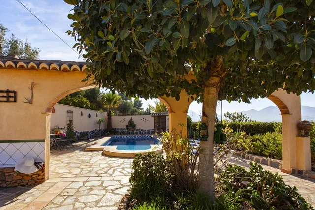 La piscina enmarcada por los arcos del jardín, con un ficus de gran porte dando sombra y la sierra asomando por la derecha