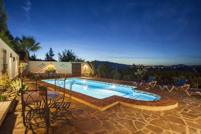 La piscina iluminada al anochecer, con el agua turquesa, mesa de forja en el solárium y las luces del valle recortadas al fondo
