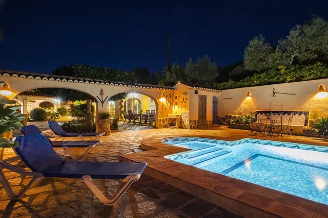 La piscina de noche, con los arcos del porche iluminados en tonos cálidos y las tumbonas sobre el solado de piedra irregular