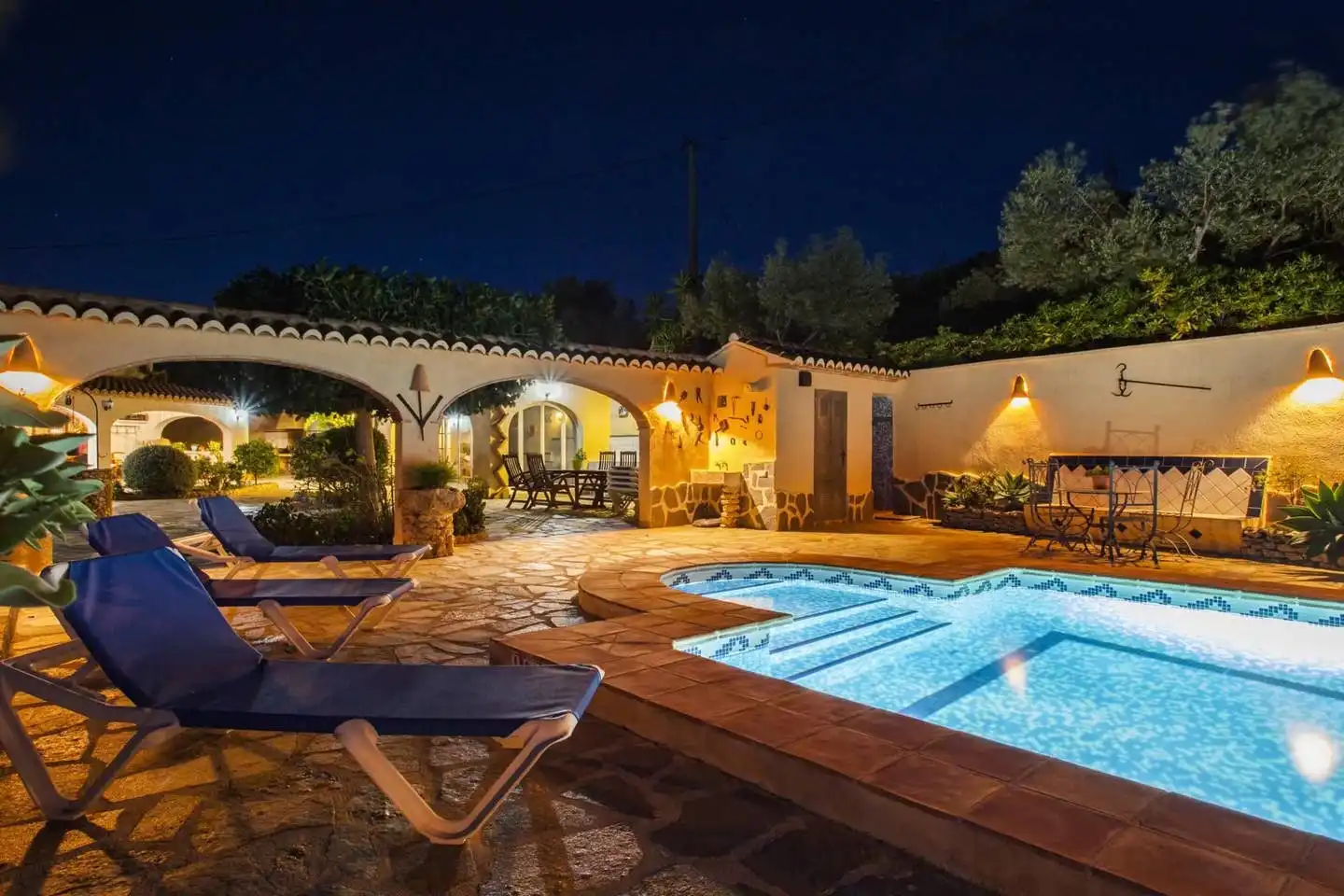 La piscina de noche, con los arcos del porche iluminados en tonos cálidos y las tumbonas sobre el solado de piedra irregular