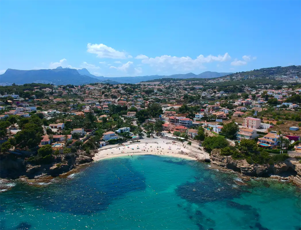 Vista aérea de una cala de arena entre rocas bajas, con el agua turquesa en primer plano, casas bajando hasta la orilla y la sierra al fondo