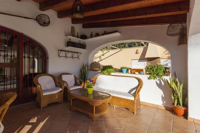 Zona de estar del porche con sofá y butacas de mimbre tapizados en blanco, junto al ventanal que asoma a la barbacoa