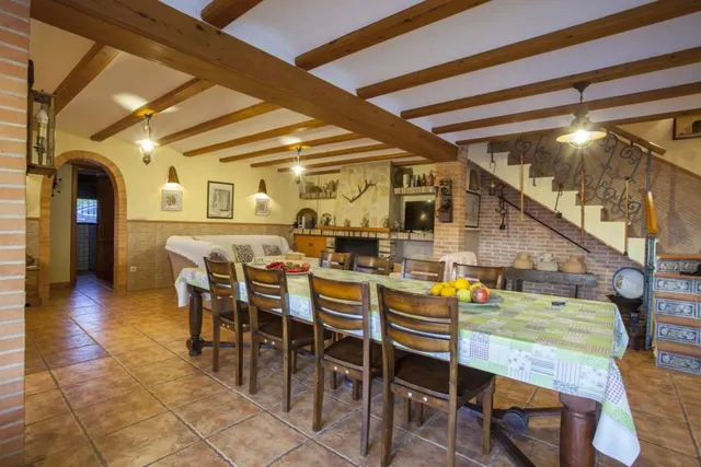 Salón de planta abierta con chimenea de obra y televisión de pantalla plana, junto a la mesa larga del comedor y la escalera de forja