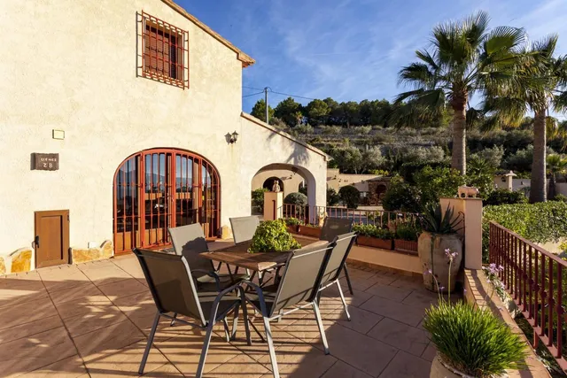Terraza junto a la fachada de la casa, con mesa para cuatro, palmeras y la ladera de pinos detrás