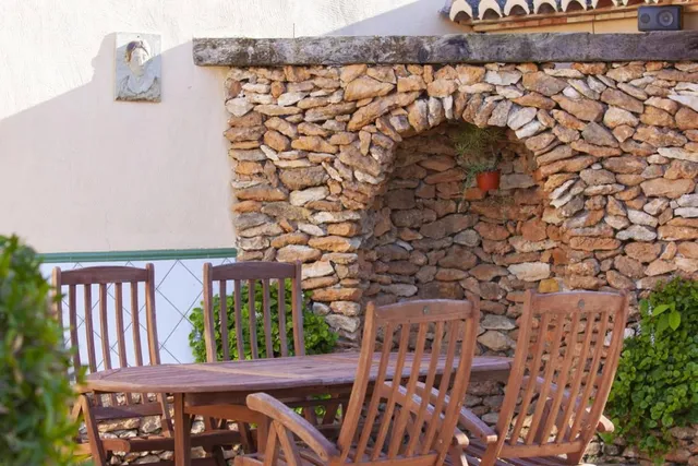 Mesa de madera para seis en el patio, delante de un muro de piedra seca rematado con una hornacina